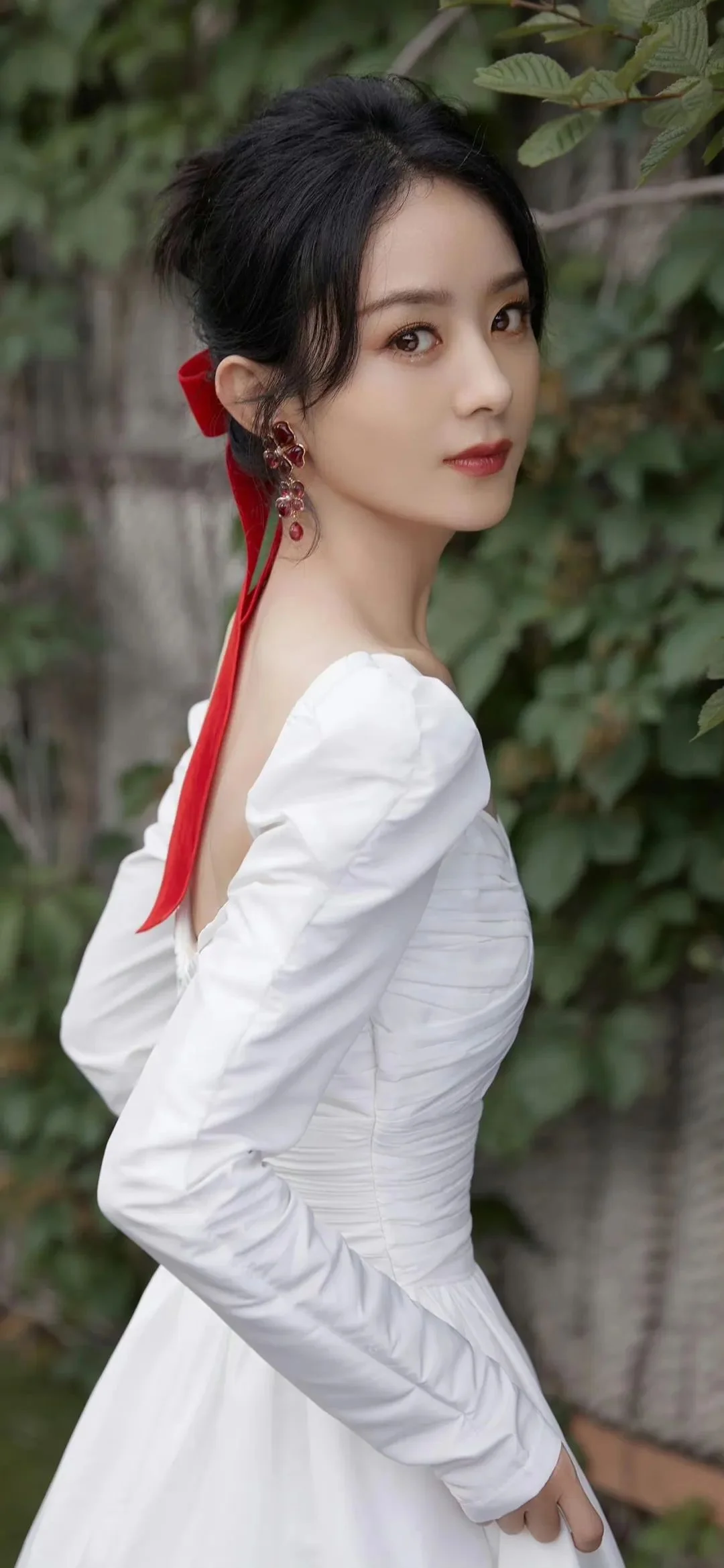 흰색 긴소매 원피스와 빨간 리본을 착용한 성인 여성이 푸른 잎사귀를 배경으로 뒤돌아 카메라를 바라보고 있다.