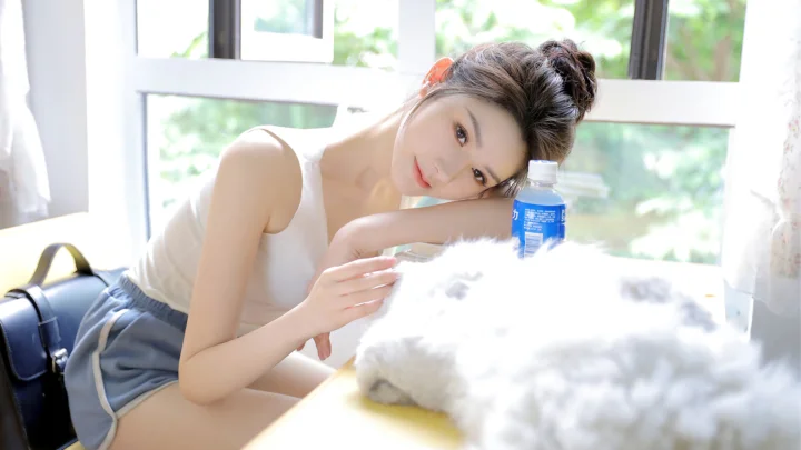 밝은 창가에 성인 여성이 기대어 있고 테이블 위에는 파란 병과 흰색의 복슬복슬한 물체가 놓여 있습니다.