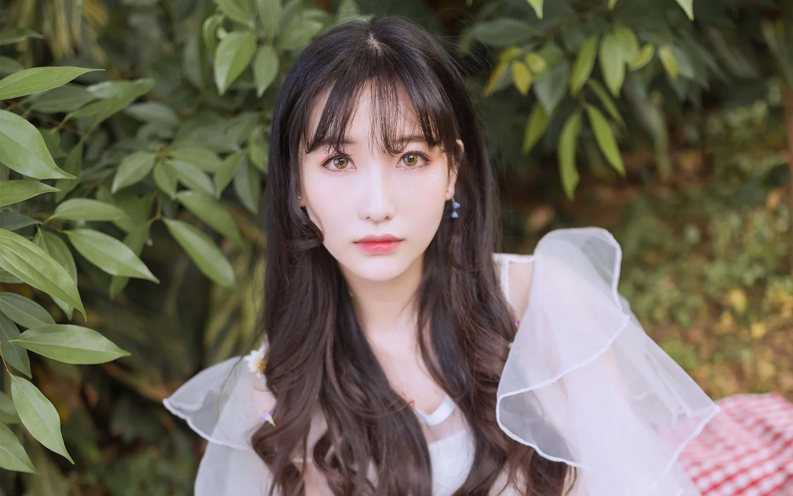 一名长发女子站在绿叶环绕的户外环境中，身穿白色轻纱上衣。