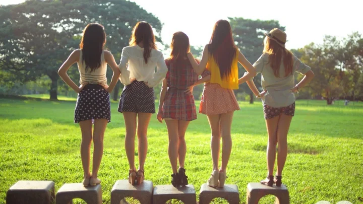 다섯 명의 여성이 잔디 공원에서 낮은 돌 받침대 위에 등을 보인 채 햇빛을 받고 서 있습니다.