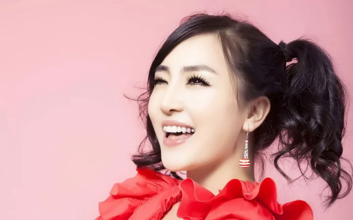분홍색 배경 앞에서 빨간색 러플 상의를 입은 양갈래 머리의 성인 여성이 미소 짓고 있다.