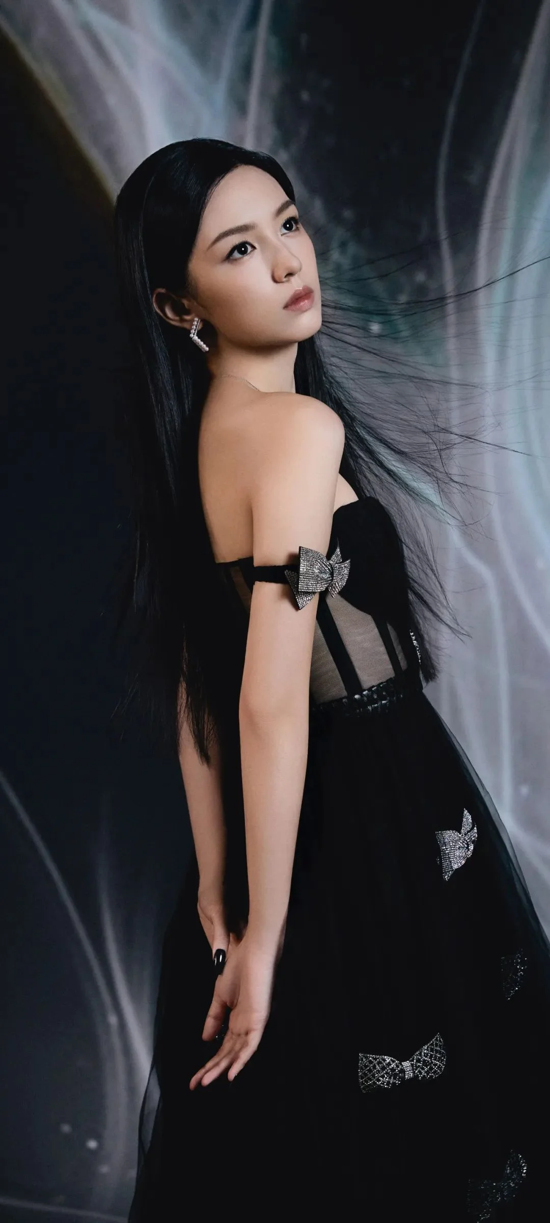 검은 드레스를 입은 여성이 어두운 배경 앞에서 뒤돌아 카메라를 바라보고 있다.