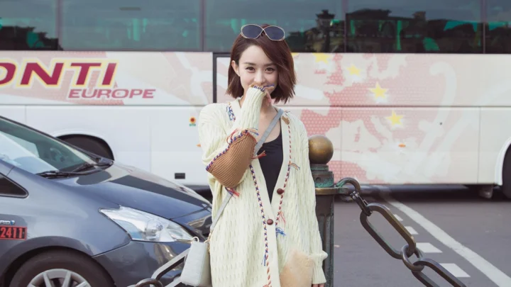 一名戴着太阳镜的女子站在城市街道旁，背景可见汽车和公交车。