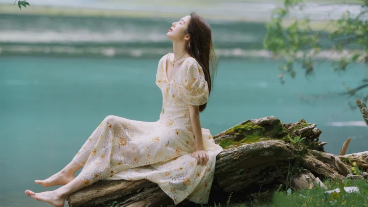 一名身穿浅色花裙的女子坐在湖畔的树干上，背景是碧绿色湖水与树木。