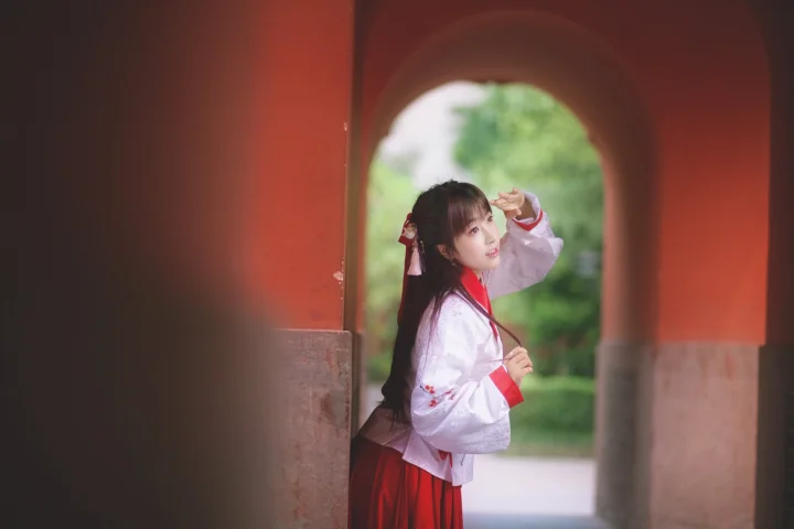전통 의상을 입은 여성이 붉은 아치 옆에 서 있고 뒤로 흐릿한 녹색 식물이 보입니다.