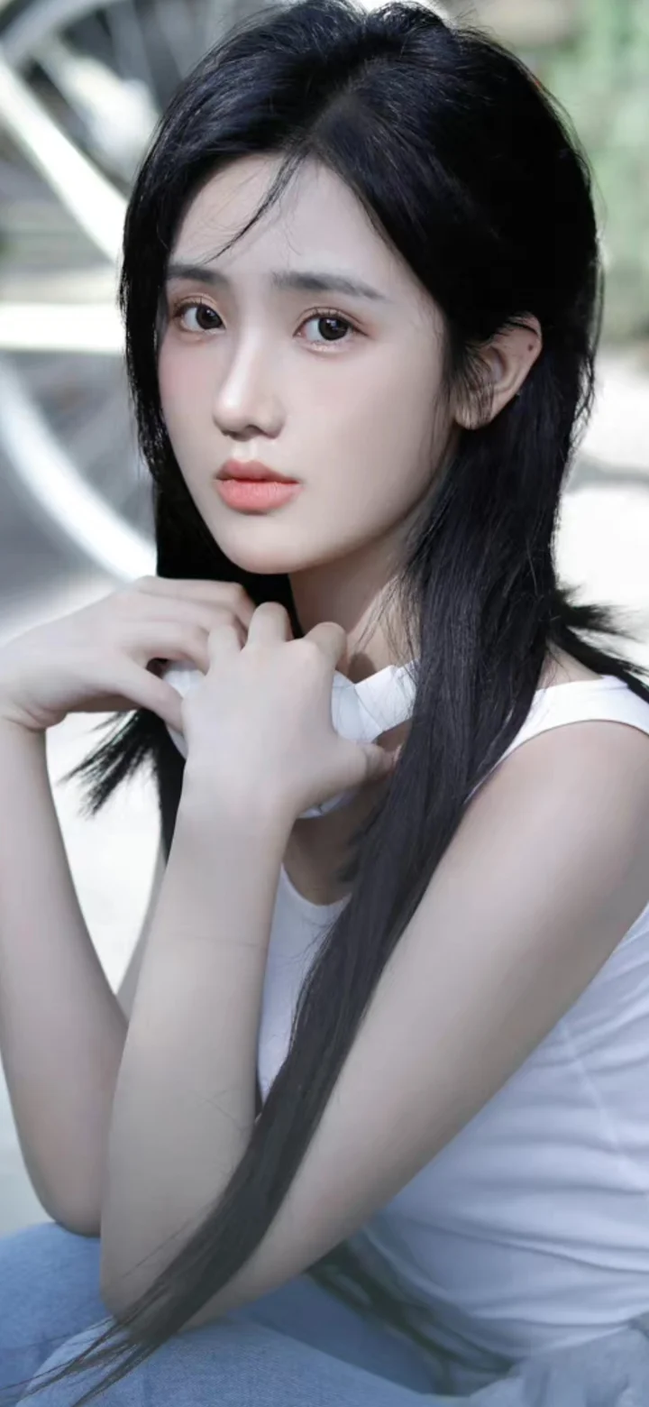 검은 긴 머리의 여성이 흰색 민소매 상의를 입고 목 근처에 손을 대며 카메라를 향해 포즈를 취하고 있다.