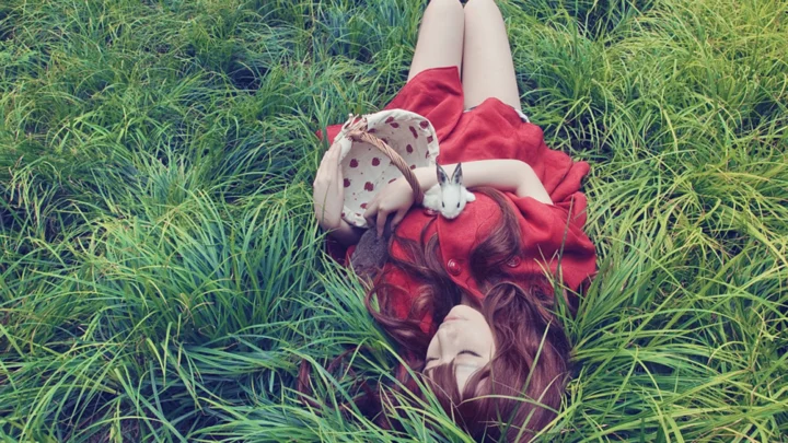 一名身穿红裙的女子仰卧在茂密的绿草中，身旁放着一只白色兔子玩偶。