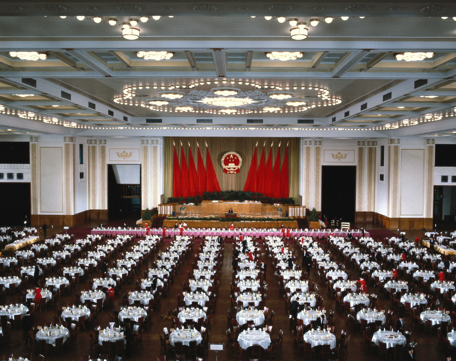 大型宴会厅内设有装饰华丽的主席台、成排的圆桌、红旗和 ornate ceiling lights。