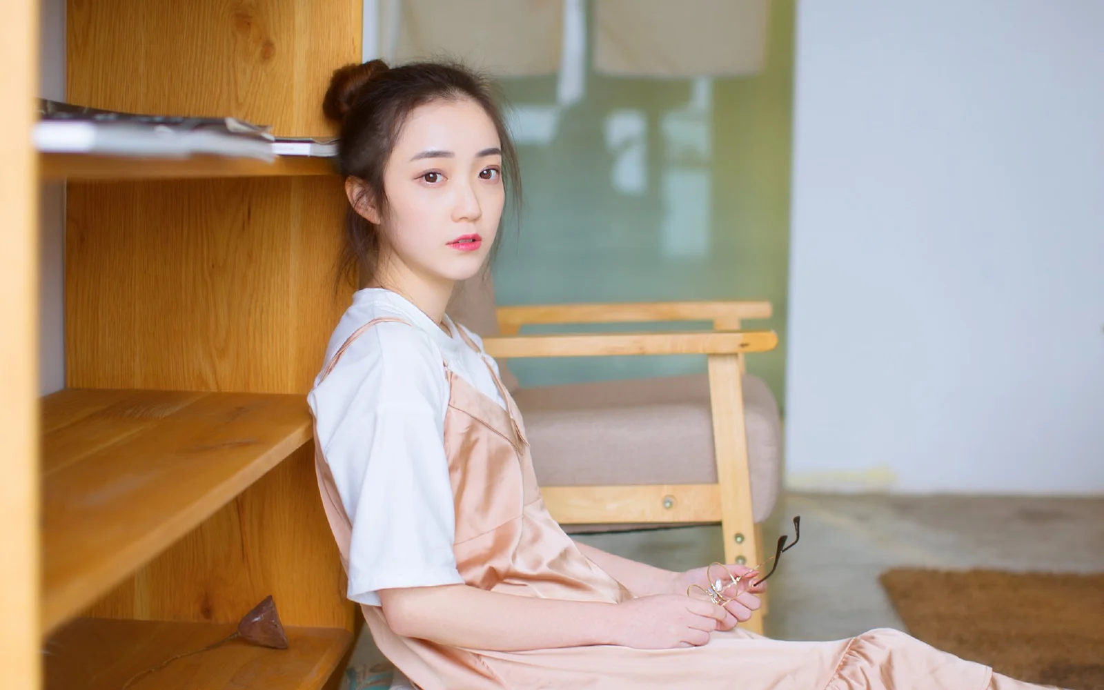 成人女性が室内の木製棚のそばに座り、眼鏡を手にしてカメラを見つめています。