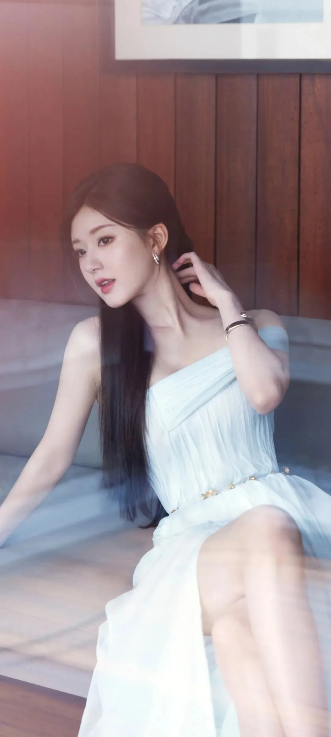 긴 머리의 여성이 흰색 드레스를 입고 실내 소파에 앉아 있습니다.