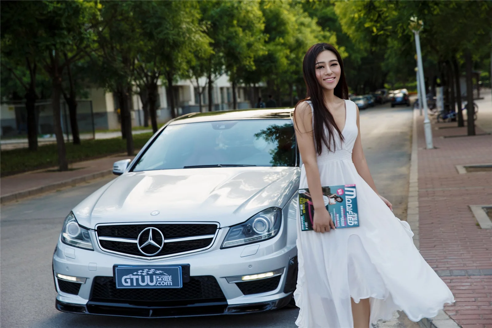 一名成年女性站在白色轎車旁，背景是一條綠樹成蔭的城市街道。