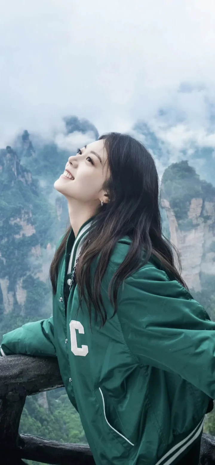 一名穿绿色夹克的女子站在云雾缭绕的山景前仰头微笑。