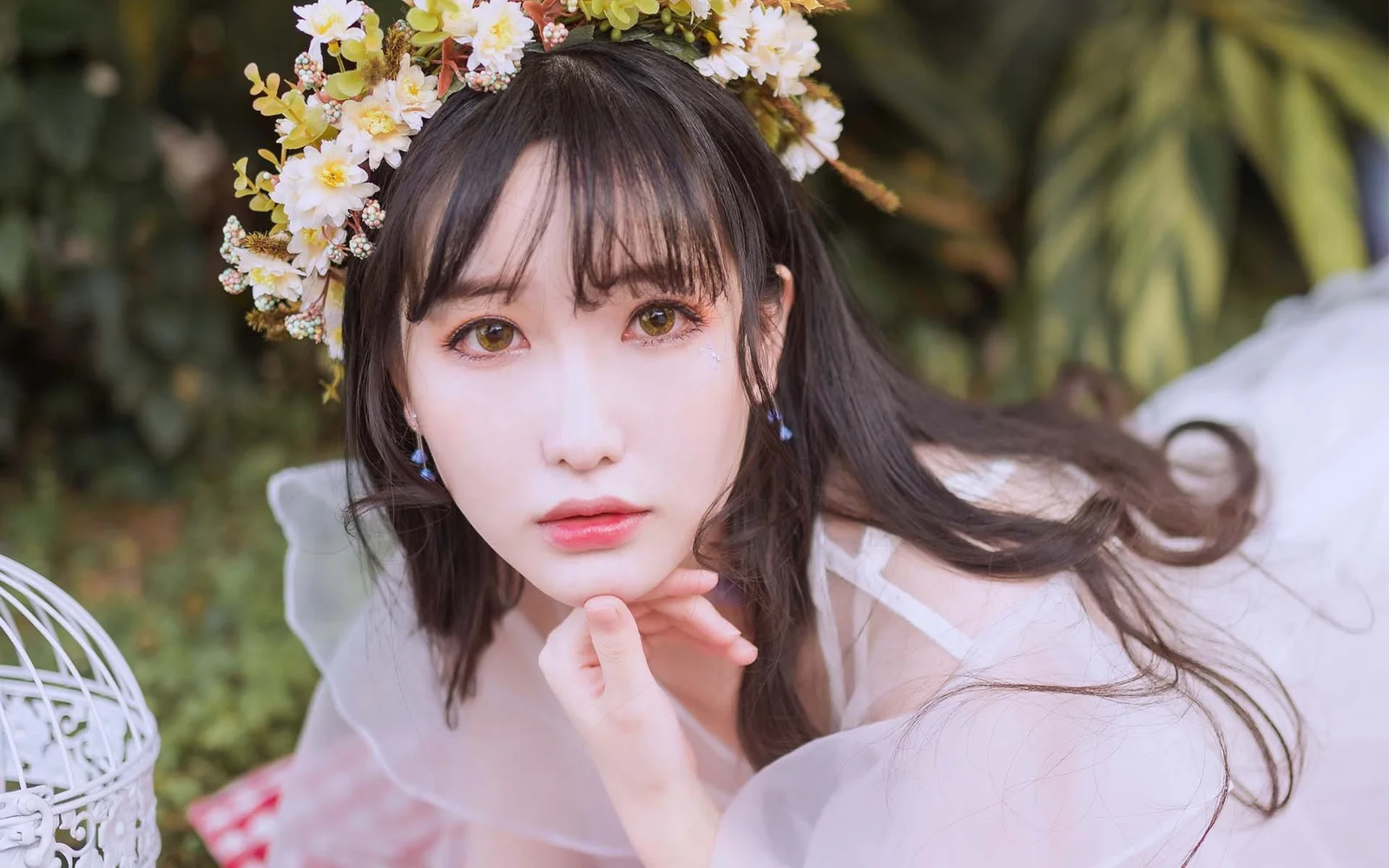 一位戴著花環、留著長髮的女性在綠植背景前看向鏡頭。
