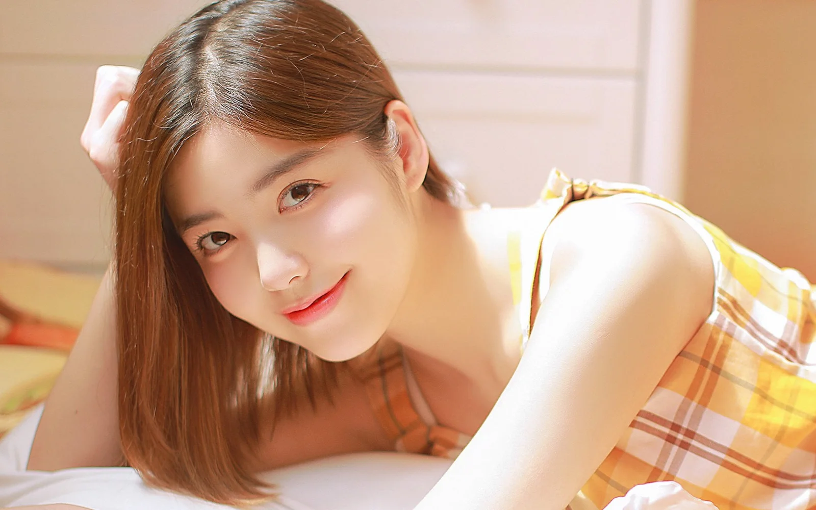 一名棕色长发女性在室内暖光下靠近镜头微笑，身穿黄色格纹无袖裙。