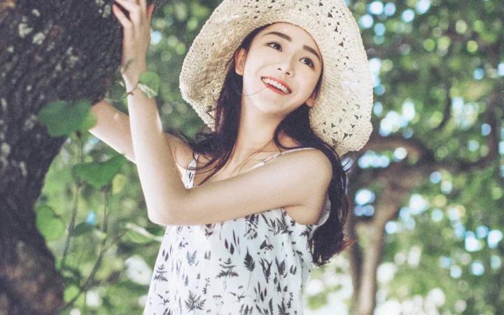 一名戴草帽的成年女子站在林间，面带微笑，身穿印花无袖上衣。