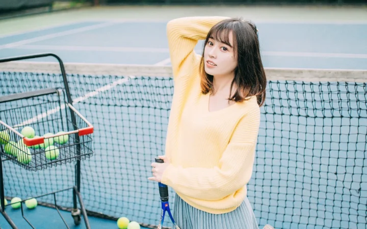 一名穿黄色针织衫和百褶裙的成年女性站在网球场旁，手持网球拍，旁边放着装有网球的球车。