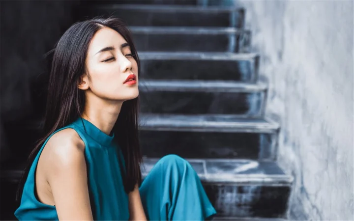 一名身穿蓝色连衣裙的成年女子坐在楼梯旁闭目侧望。