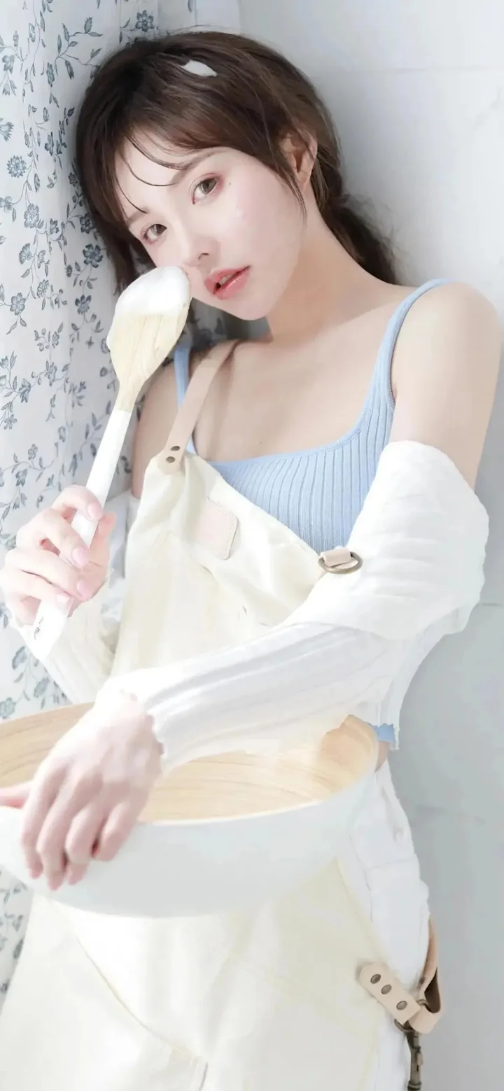 연한 파란색 상의와 흰색 앞치마를 입은 성인 여성이 실내 벽 옆에서 흰색 숟가락을 들고 카메라를 바라보고 있다.