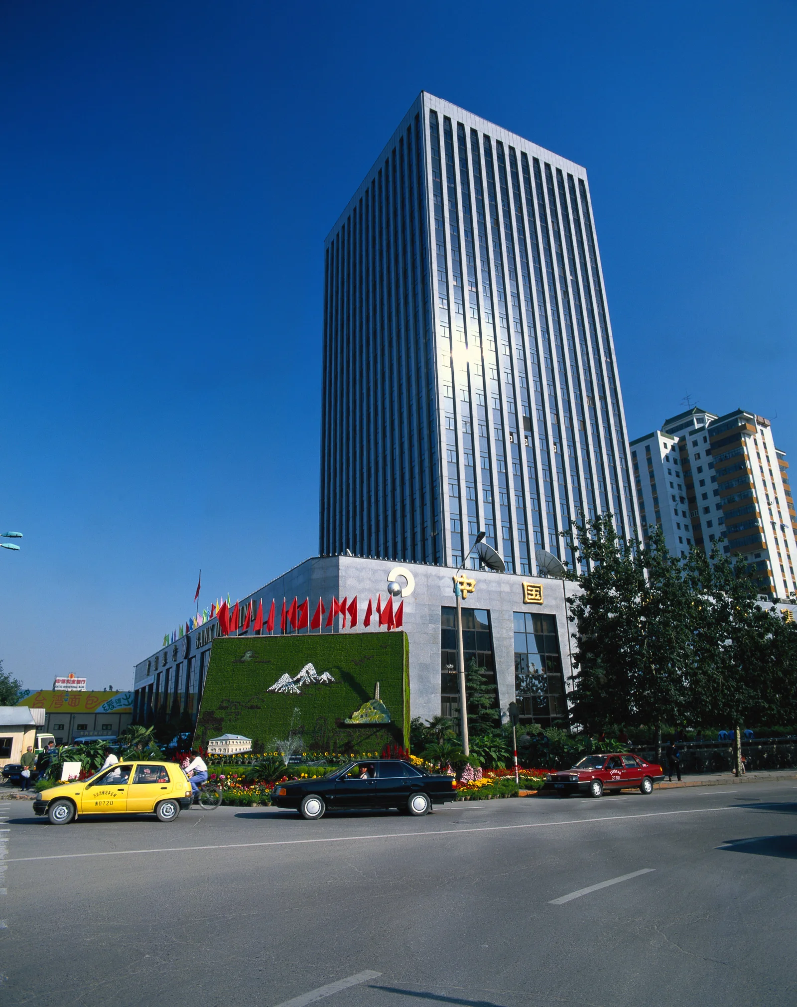 蓝天下，一座带有旗帜和绿化装饰的高层建筑临街而立，前方道路上有车辆经过。