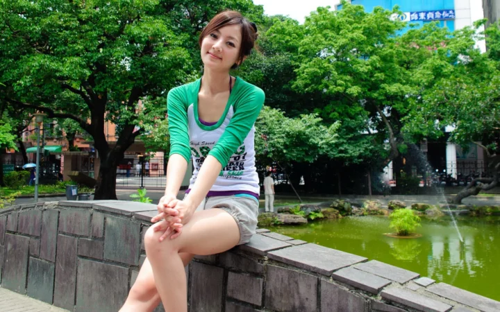 一名成年女性坐在公园池塘边的石墙上，周围可见绿树和水面。