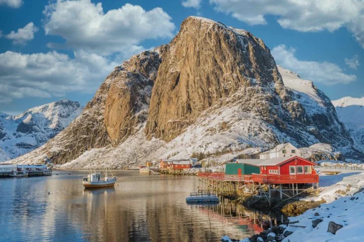 一座积雪覆盖的陡峭山峰矗立在宁静的峡湾旁，岸边点缀着红色房屋和一艘小船。