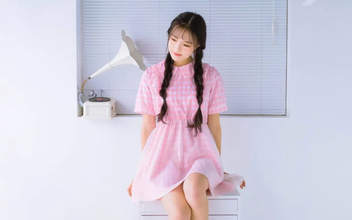 분홍색 원피스를 입고 땋은 머리를 한 여성이 실내 창가에 앉아 있습니다.