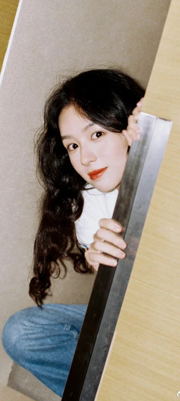 긴 머리의 여성이 실내 문 뒤에서 몸을 내밀고 카메라를 바라보며 미소 짓고 있다.
