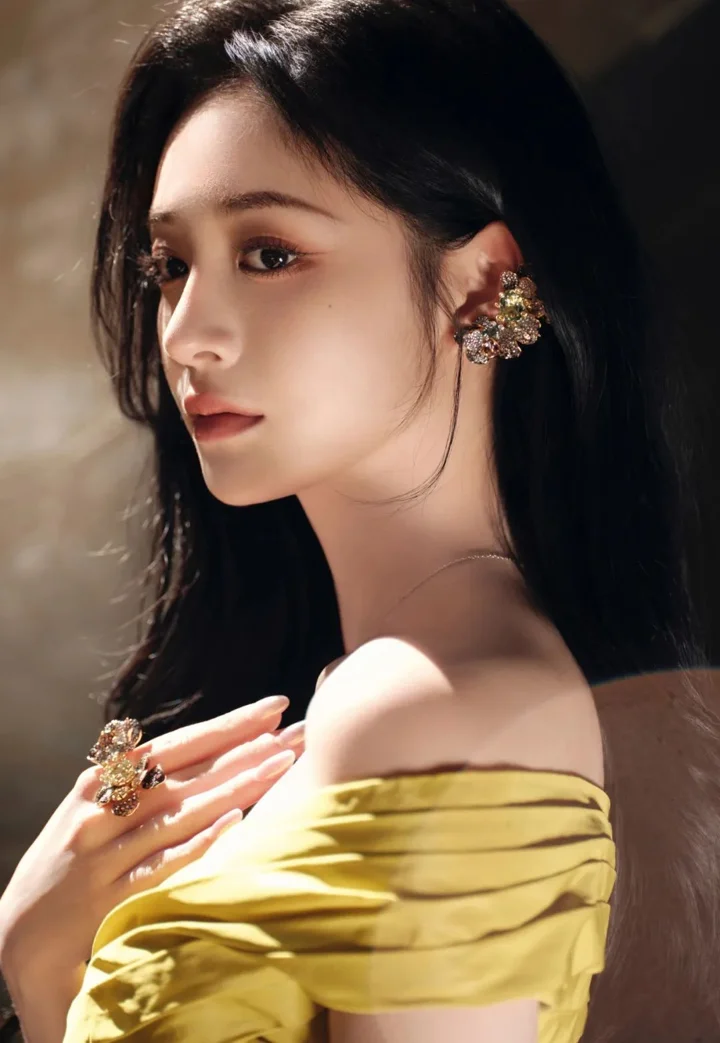 긴 검은 머리의 여성이 노란색 오프숄더 상의를 입고 부드러운 조명 아래에서 옆모습을 보이는 인물 사진입니다.