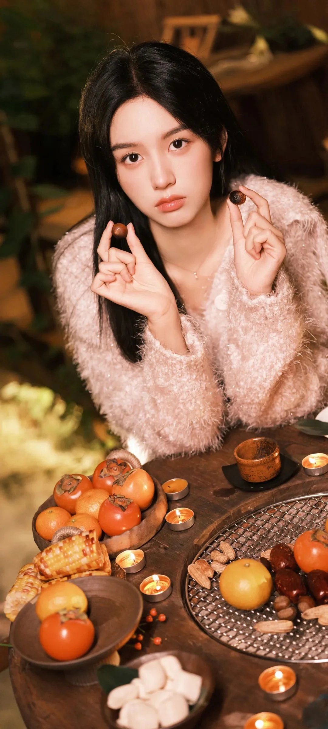 黒髪の女性が淡いピンクのセーターを着て、食べ物が並んだテーブルのそばに座っています。