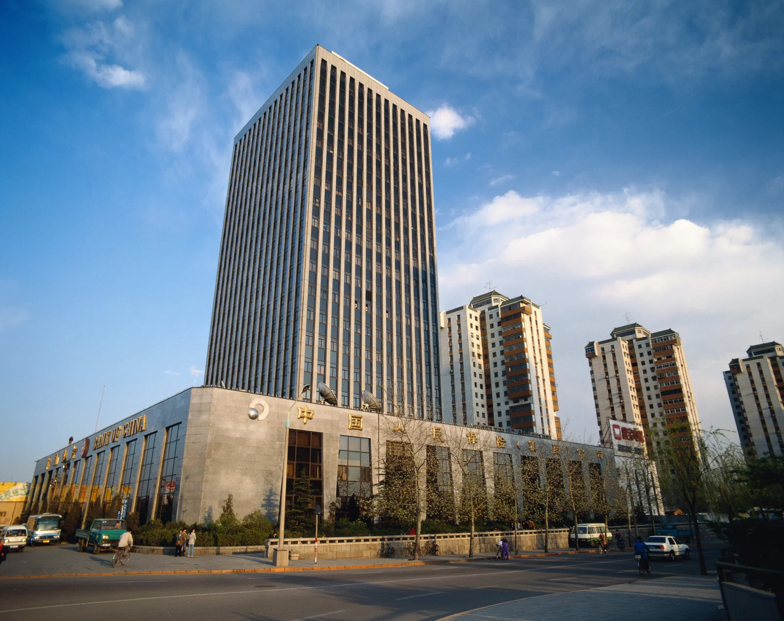 一座高耸的城市建筑矗立在蓝天和白云下，前方是街道与低层建筑。