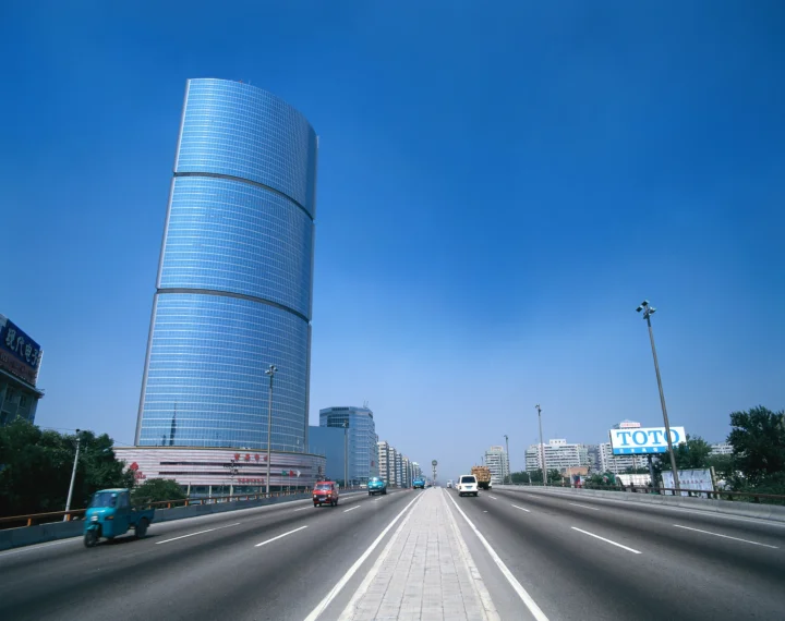 宽阔的城市高速公路通向远方，旁边矗立着弧形玻璃高楼，天空晴朗湛蓝。