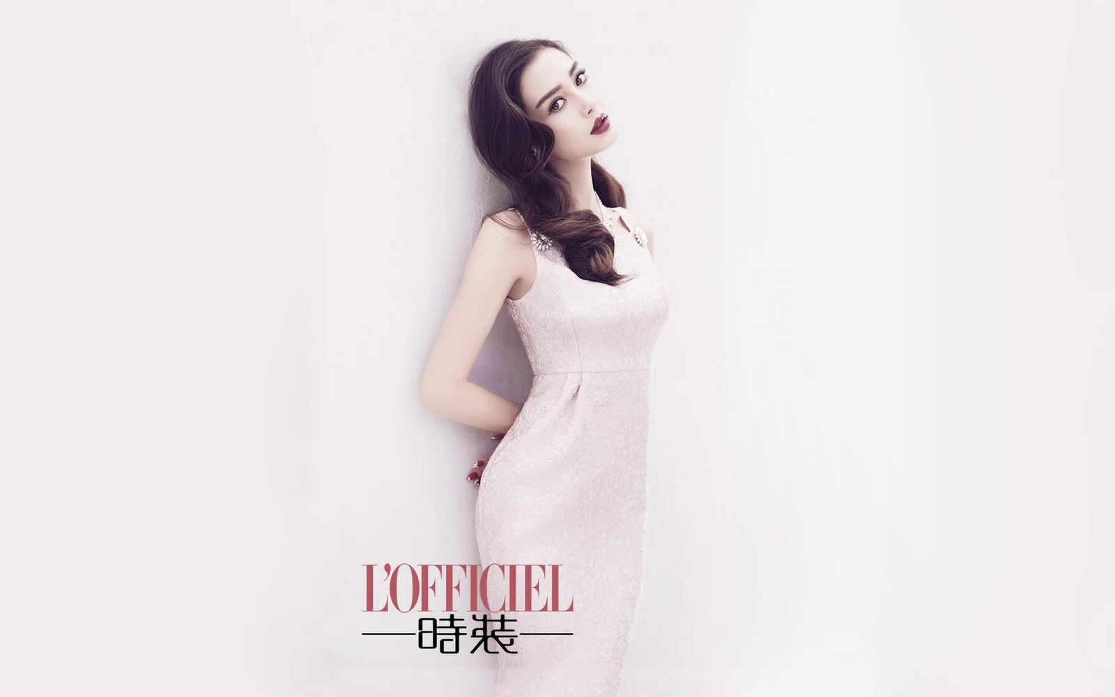 一位成年女性身穿浅色无袖连衣裙靠墙站立，呈现简洁的时尚肖像画面。