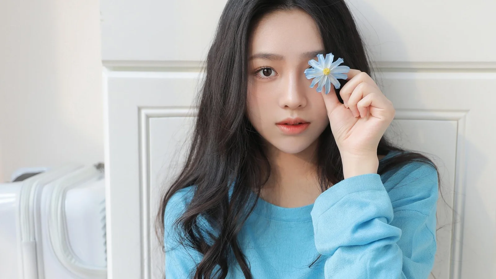 파란 스웨터를 입은 여성이 실내에서 작은 파란 꽃을 눈가에 들고 있습니다.