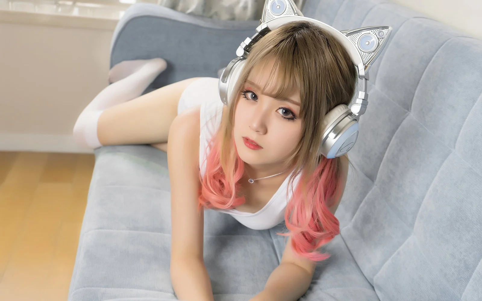 猫耳ヘッドホンを着けたピンクのグラデーションヘアの女性が、グレーのソファにうつ伏せになってカメラを見ています。