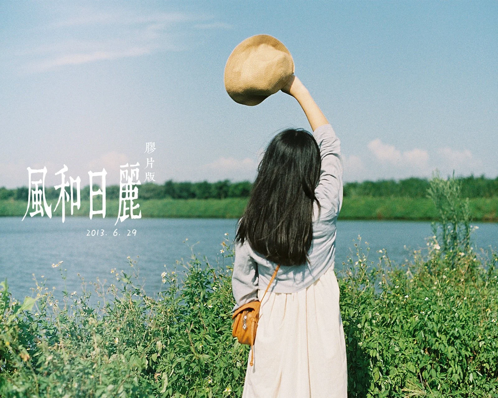 長い髪の女性が湖畔の草地に立ち、帽子を頭上に掲げて湖を眺めています。