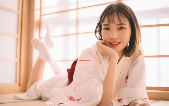 一名短发女性穿着白色衣服，趴在明亮窗边微笑看向镜头。