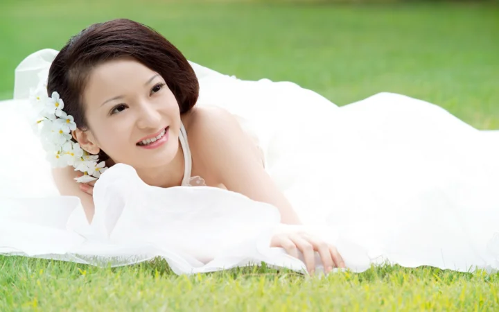 一名女子躺在草地上的白色床单上微笑，头发旁点缀着白色花朵。
