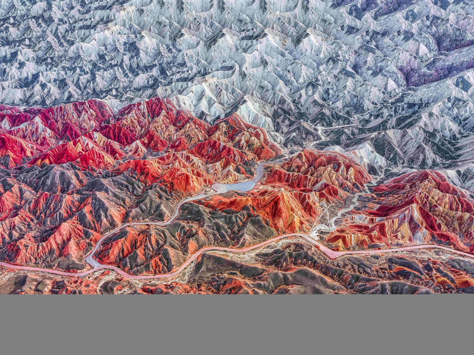 俯瞰の構図で、赤や灰色、紫色に重なる山々と、その間を通る曲がりくねった道路が見える。