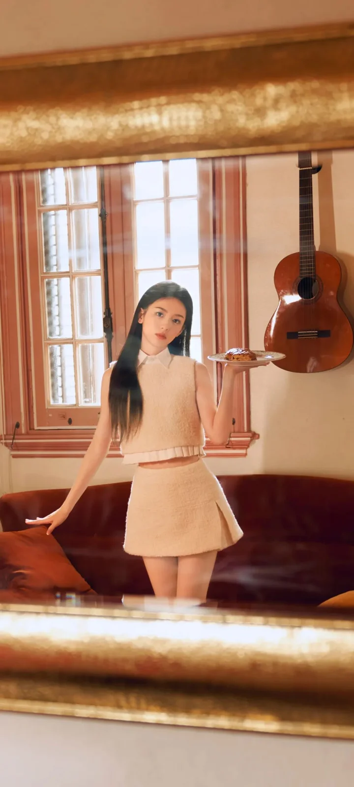 一名成年女性站在室内镜子前，手持托盘，墙上挂着一把吉他。