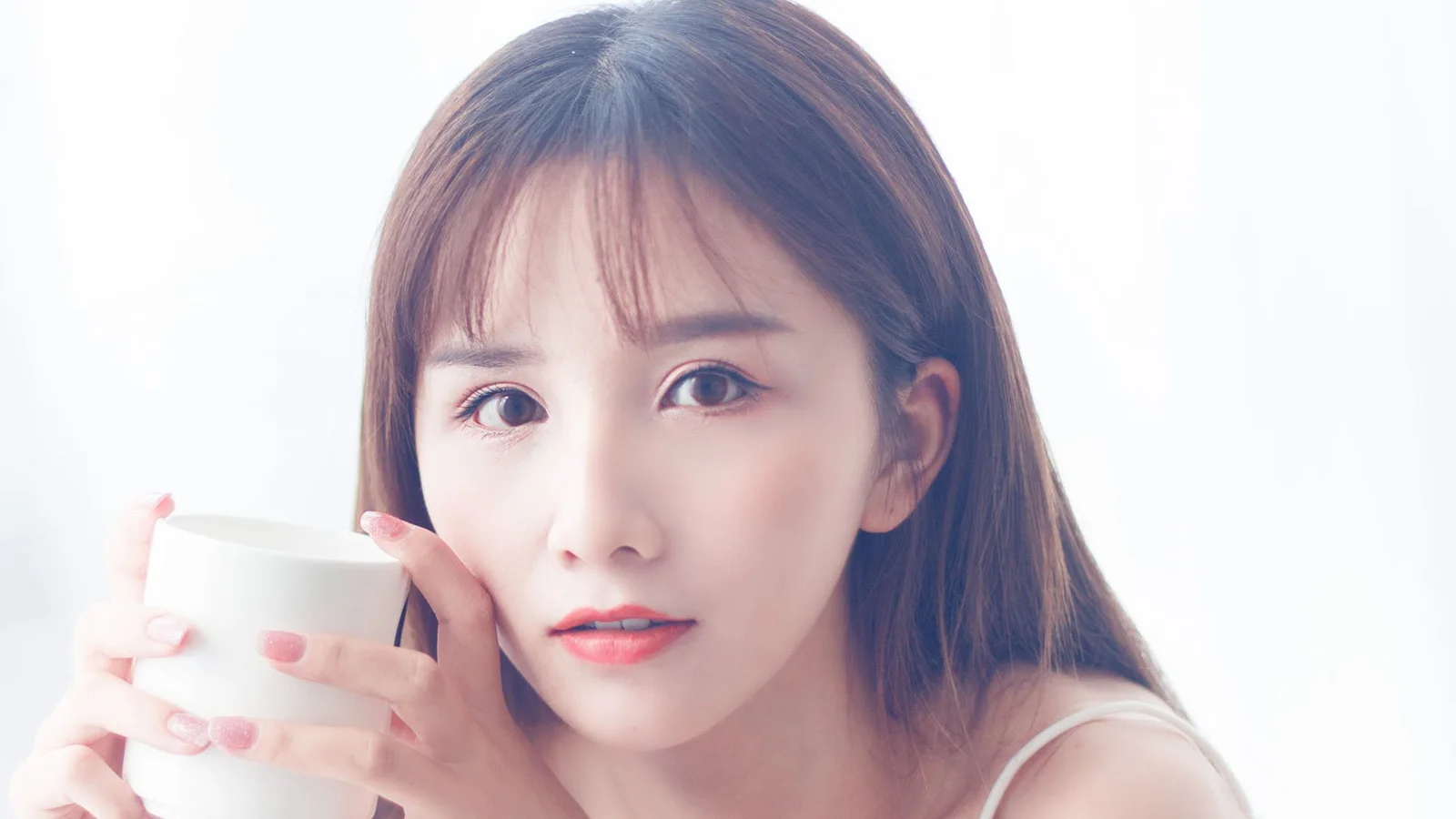 밝고 단순한 배경에서 흰색 컵을 들고 카메라를 바라보는 여성의 클로즈업 인물 사진입니다.