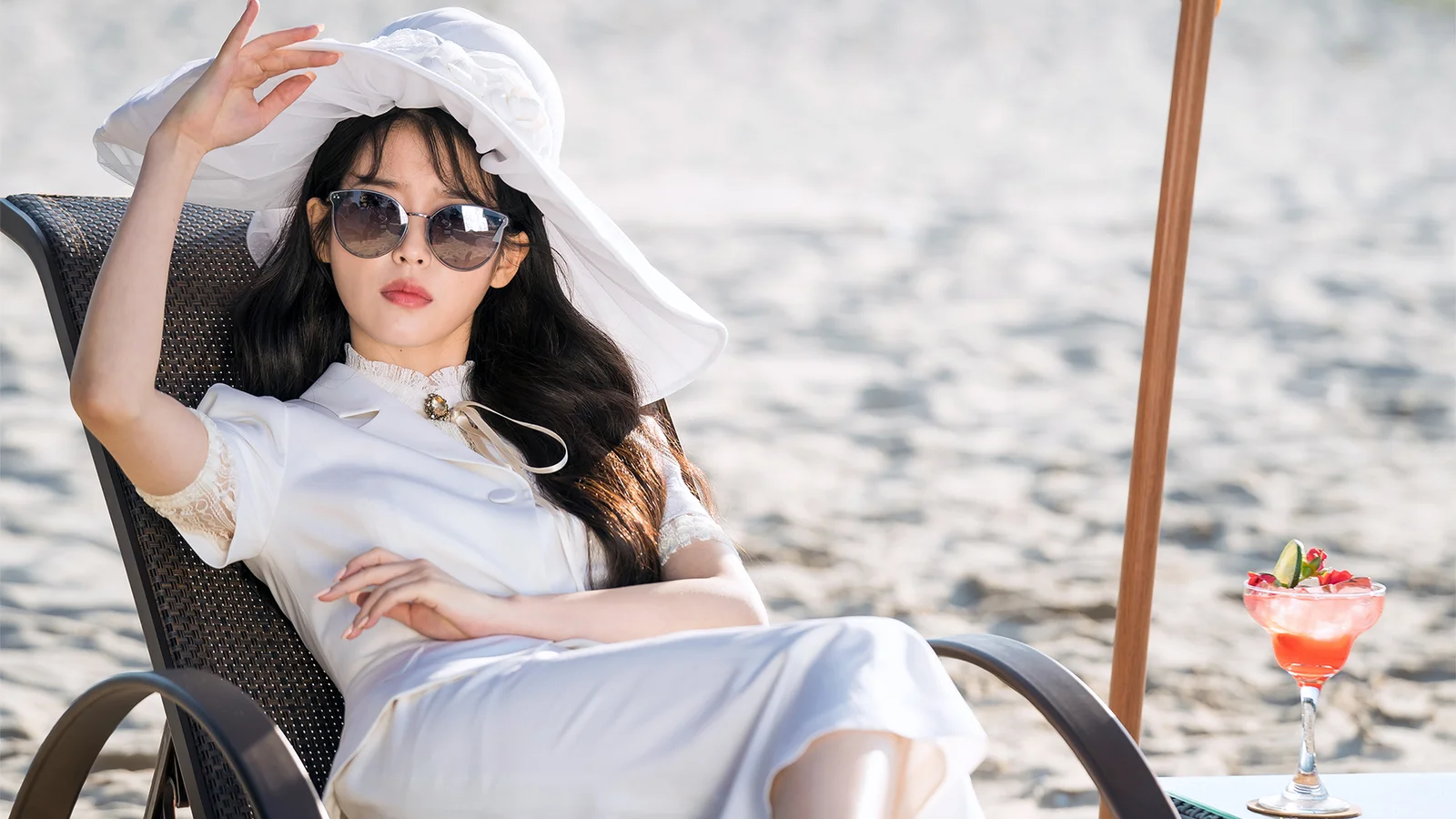 선글라스를 쓴 성인 여성이 흰색 옷을 입고 물가의 라운지 의자에 앉아 있으며 옆에 음료가 놓여 있습니다.