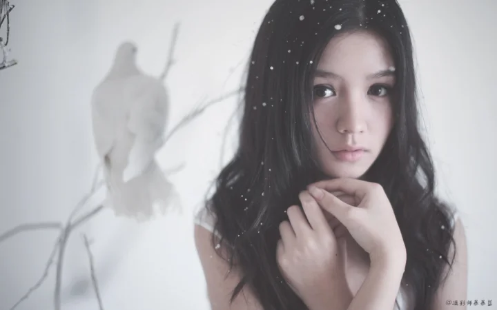 一名成年女性在浅色背景前凝视镜头，黑发周围飘落着白色雪点。