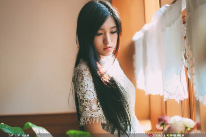 一名长发女子站在窗边，身穿蕾丝上衣，室内沐浴在温暖的阳光中。