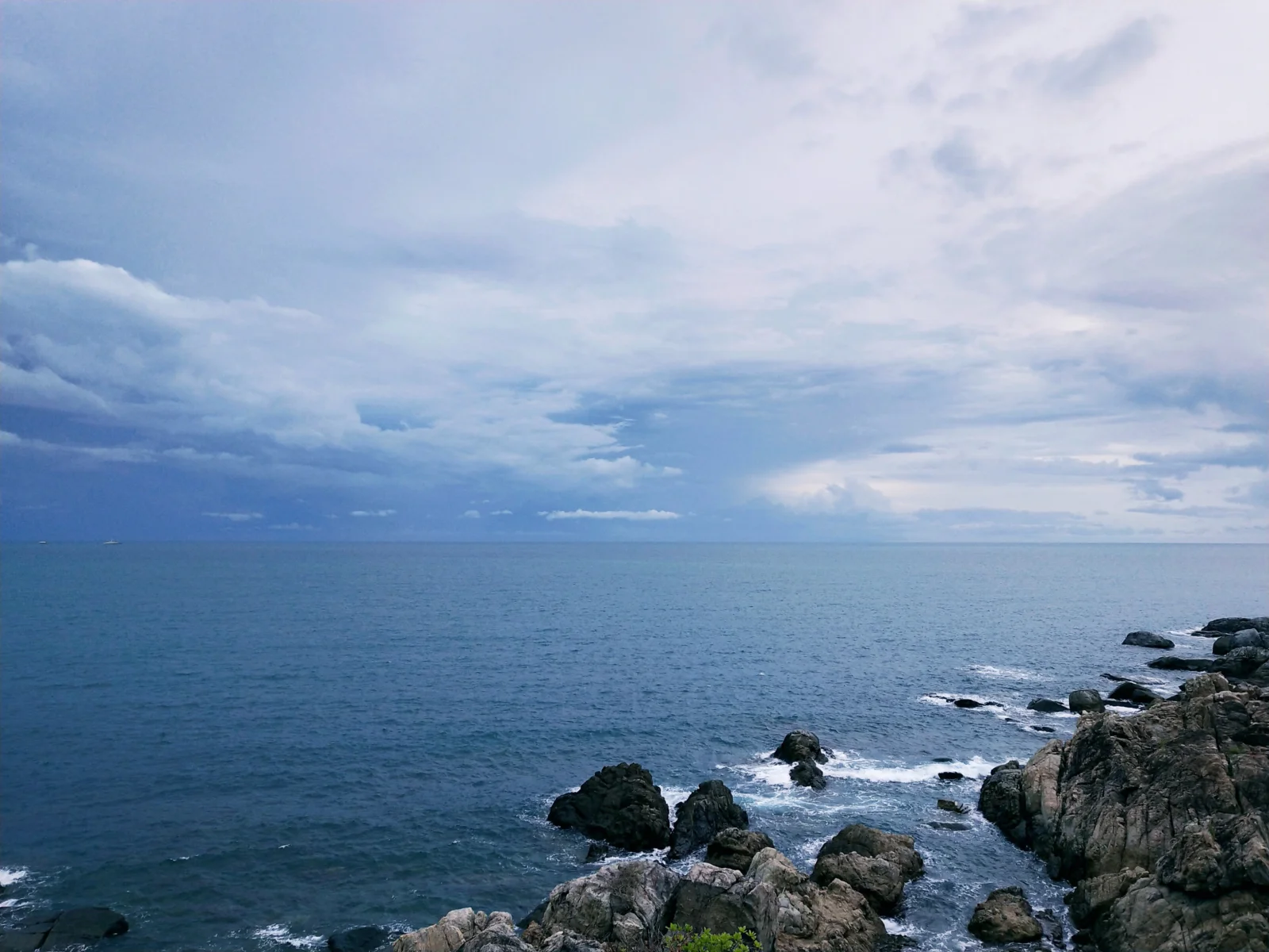 辽阔海面延伸至远方地平线，前景是被海浪拍打的岩石海岸，天空布满层叠云彩。