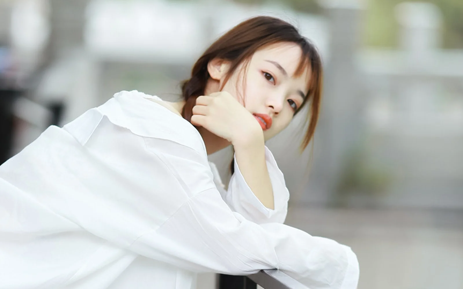 흰 셔츠를 입은 여성이 실내에서 옆으로 앉아 손으로 턱을 괴고 카메라를 바라보고 있습니다.