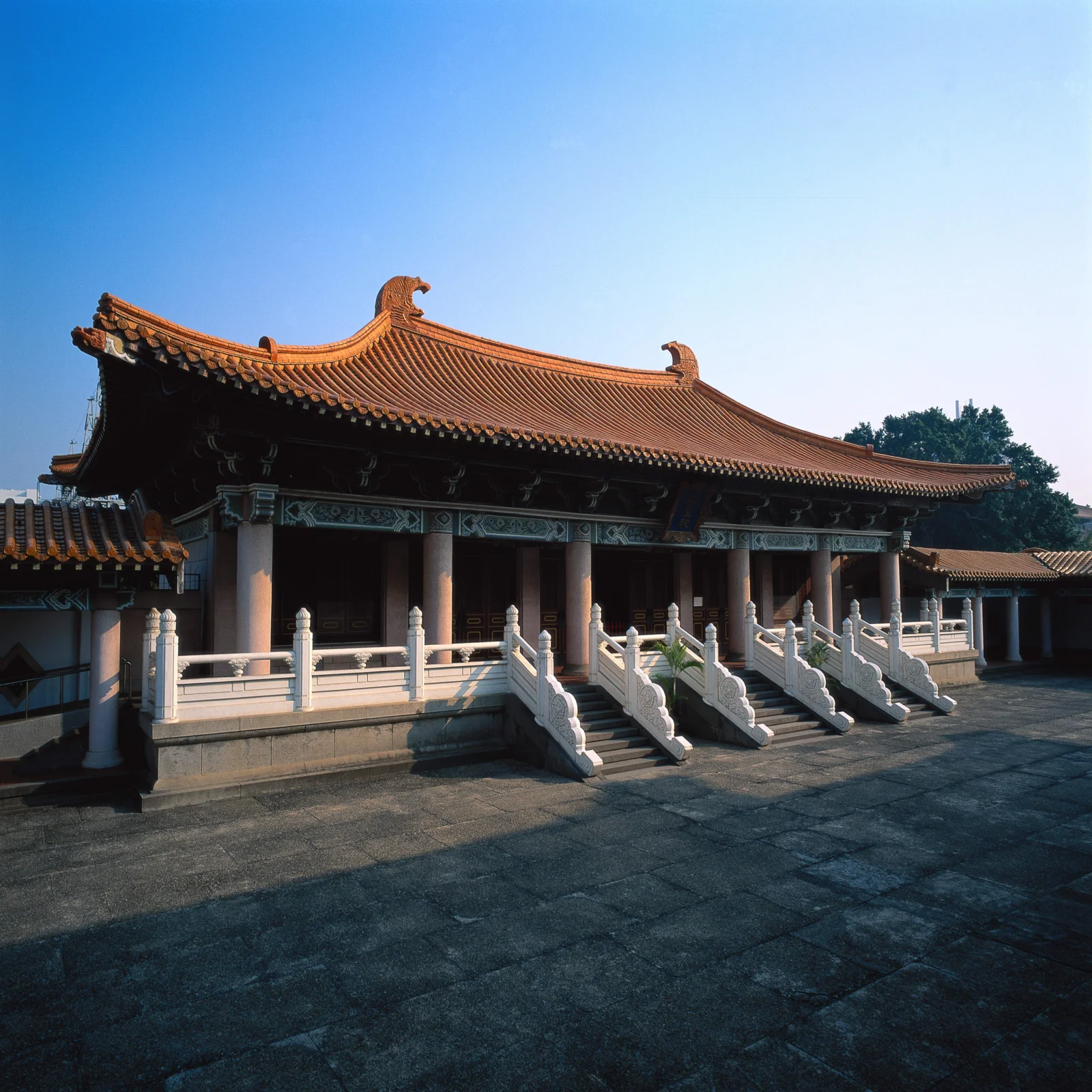 蓝天下，一座带橙色琉璃瓦屋顶和白色石栏杆的传统建筑坐落在石板庭院中。