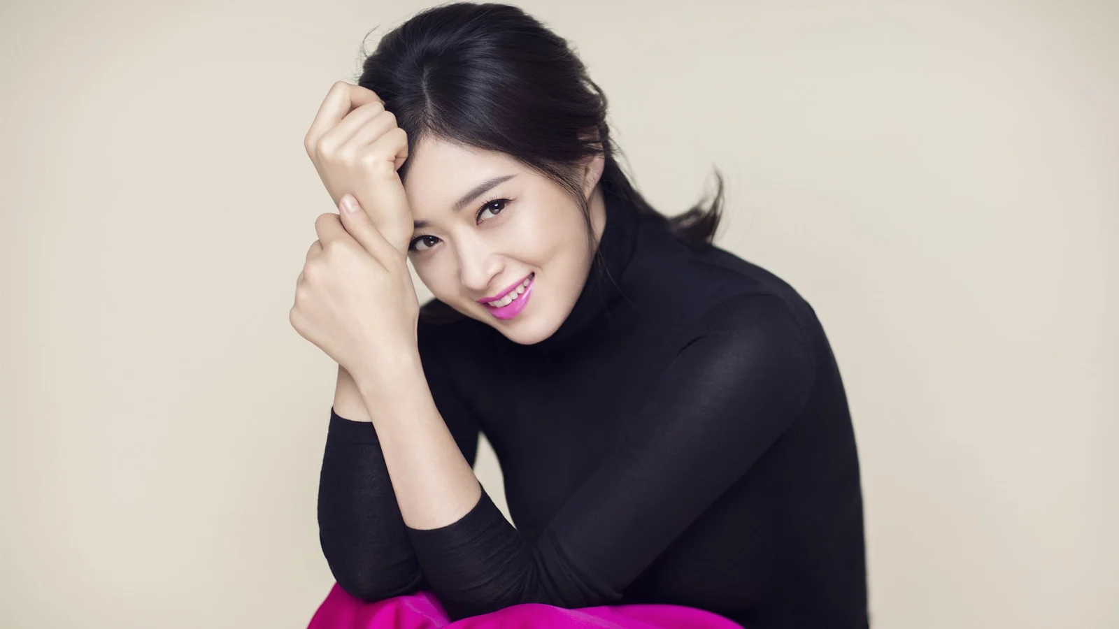 검은색 터틀넥을 입은 성인 여성이 밝은 단색 배경 앞에서 카메라 가까이 미소 짓고 있다.