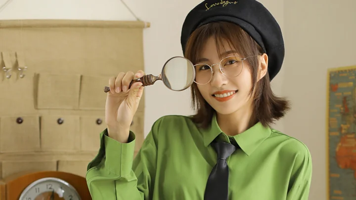 검은 베레모와 둥근 안경을 쓴 여성이 초록색 셔츠를 입고 돋보기를 들고 카메라를 향해 미소 짓고 있다.