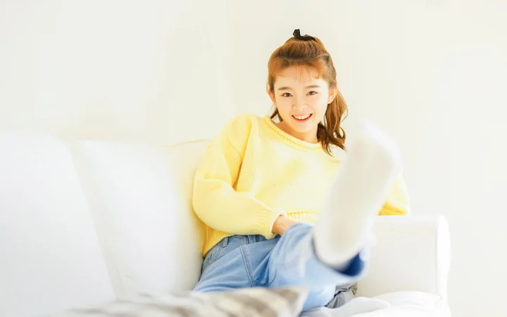 노란색 스웨터와 청바지를 입은 젊은 여성이 흰색 소파에 앉아 카메라를 향해 미소 짓고 있습니다.