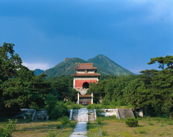 一座传统古建筑坐落在绿树环绕的山前，前方有石阶和小路，天空湛蓝。